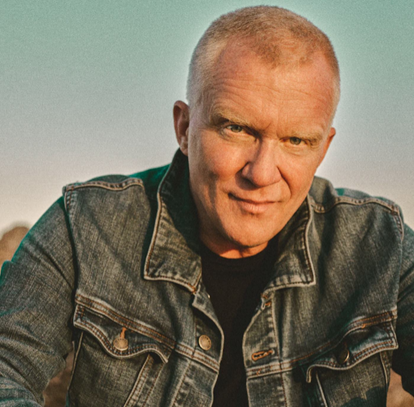 Mature man with closely cropped hair, wearing a denim jacket, smiling at the camera in a natural outdoor setting at sunset.