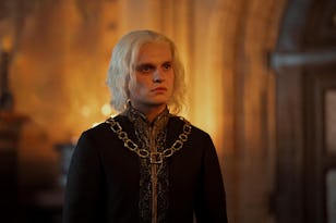 A man with long white hair and intense expression, wearing medieval-style armor, stands in a dimly lit room with a fiery glow in the background.