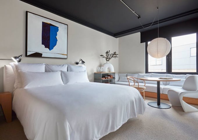Modern bedroom featuring a large white bed, black ceiling, and abstract blue art, with a bright, cozy seating area by large windows.