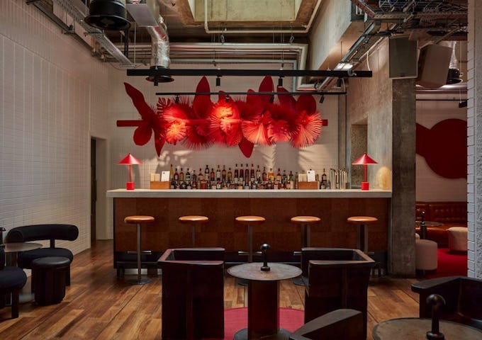 Modern bar interior with wooden floors, a well-stocked counter, striking red decor, and ambient lighting.