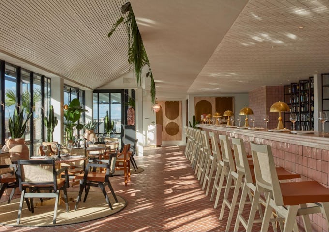 Bright and stylish restaurant interior with bar stools, dining tables, and tropical plants under a sunlit, slatted ceiling.