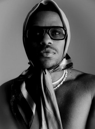 Black and white portrait of a man wearing glasses, a headscarf tied at the front, and a pearl neckla...