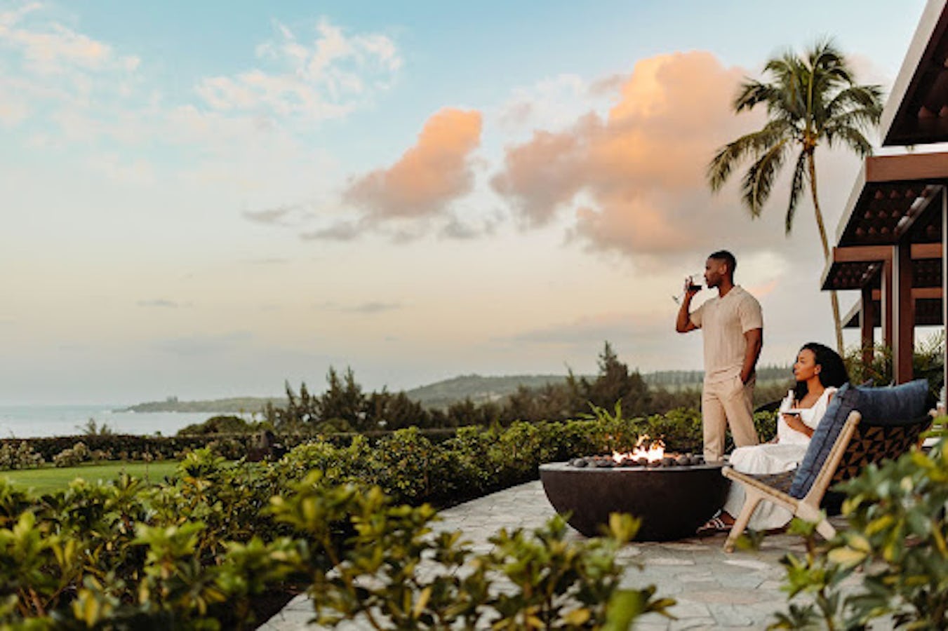 A couple enjoys a serene evening by an outdoor fire pit overlooking the ocean at a tropical resort.