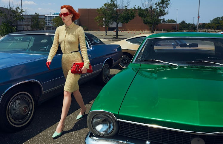 Woman with red hair in a beige dress standing between a blue and a green vintage car in a parking lo...