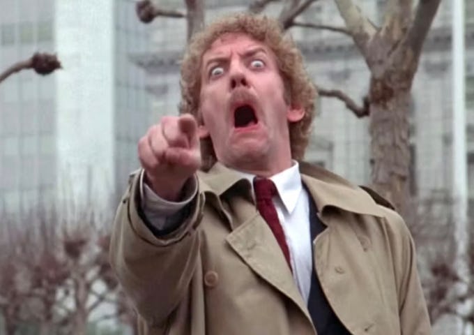 A shocked man with curly hair in a beige coat and red tie pointing and gaping in surprise, trees and a building in the background.