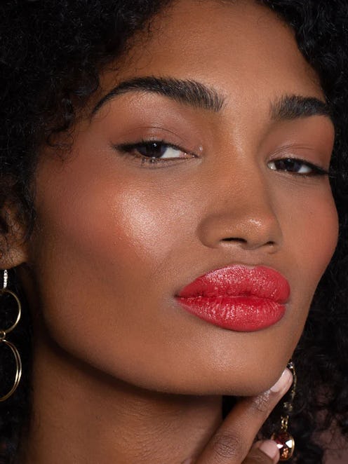 Close-up of a woman with curly hair, wearing glossy red lipstick, large earrings, and a thoughtful e...