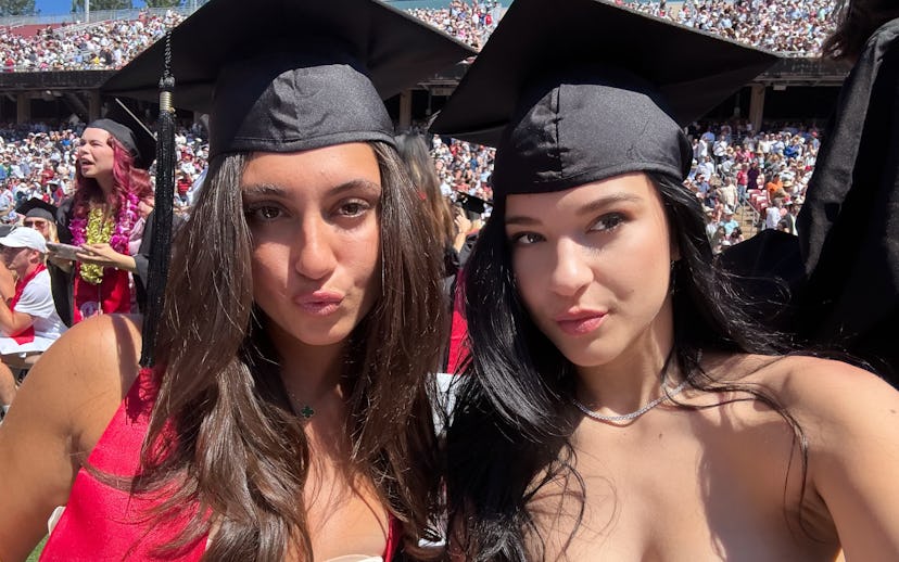 Two young women in graduation caps posing for a selfie at a crowded outdoor ceremony.