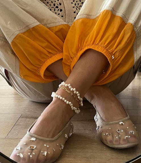 Close-up of a person's feet wearing embellished transparent shoes and pearl anklets, with vibrant or...