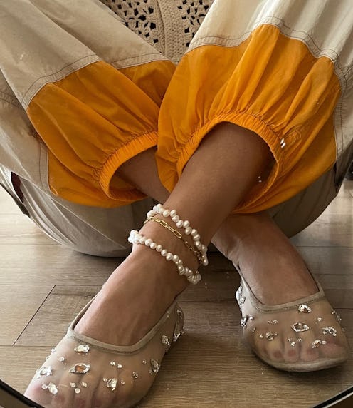Close-up of a person's feet wearing embellished transparent shoes and pearl anklets, with vibrant or...