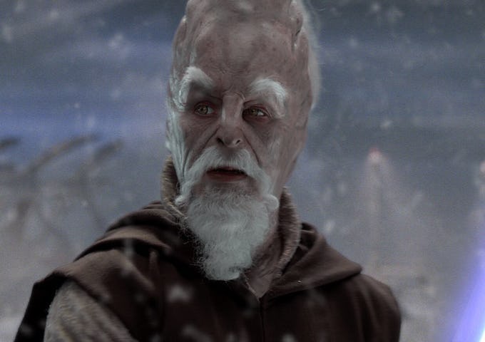An elderly alien with red skin, white beard, and intense eyes, wearing a brown robe in a snowy backdrop.
