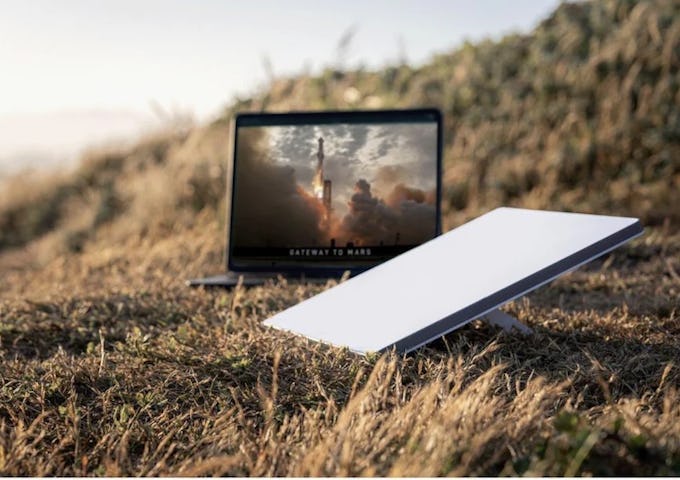 A laptop displaying a space launch image is set on grassy terrain next to a closed white book, evoking remote, outdoor work or study.
