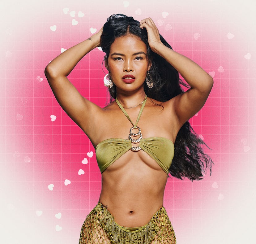 Woman in green top and skirt poses with hands on her head against a pink, heart-speckled background.