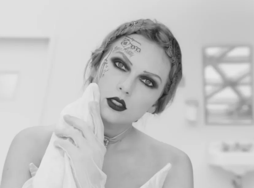 A black and white photo of a woman with intense makeup and tattoos, holding a cloth near her face in...