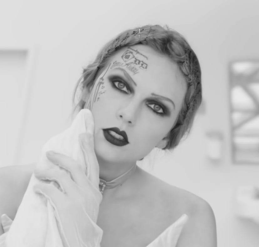 A black and white photo of a woman with intense makeup and tattoos, holding a cloth near her face in...