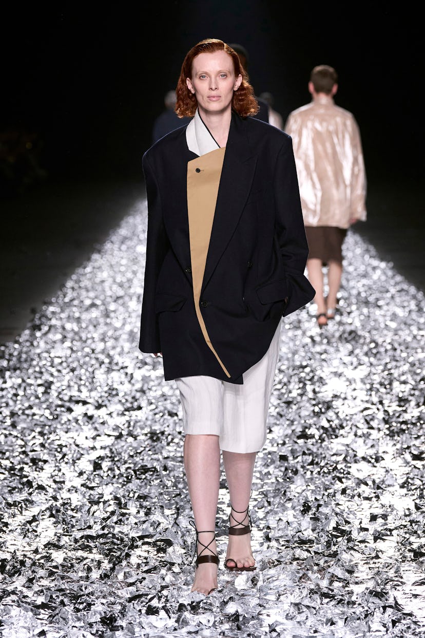 Model in oversized black blazer and beige dress walking on metallic confetti runway.