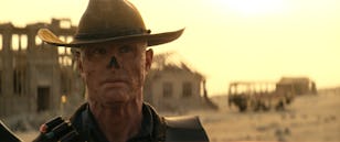 Close-up of a man in a cowboy hat with war paint on his face, against a desert landscape with dilapidated buildings.