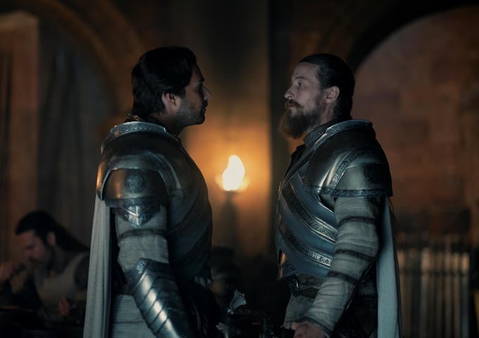 Two knights in armor converse intently inside a dimly lit medieval hall, with a lit torch on the wall behind them.