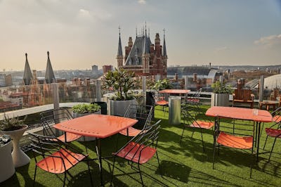 Rooftop garden with orange tables and chairs, overlooking a historic building and cityscape under a clear sky.