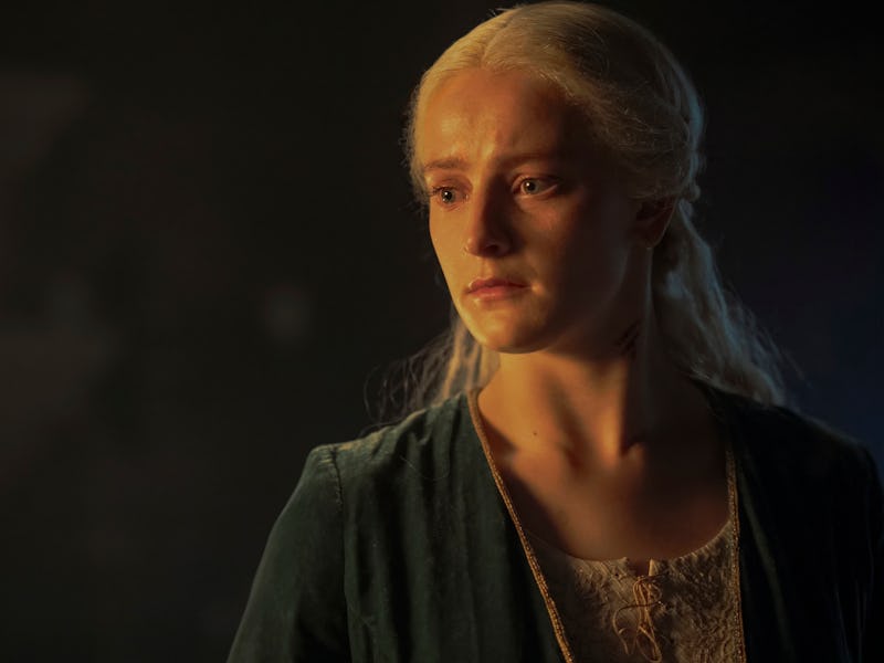 A woman with platinum blonde hair and solemn expression, wearing a green shawl over a cream dress, l...