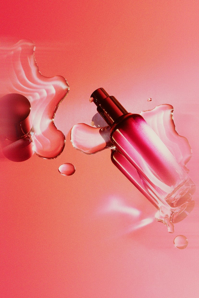 A pink skincare serum bottle on a reflective surface with scattered liquid droplets around it.