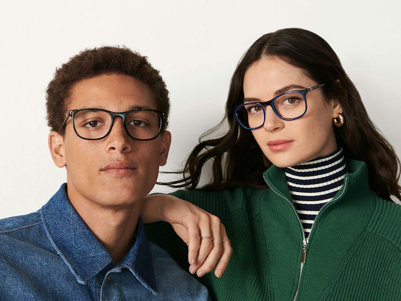 A young man and woman wearing stylish eyeglasses pose together against a plain background. The man dons a denim shirt; the woman sports a green jacket.