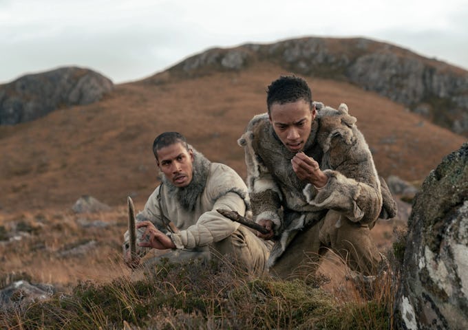 Two men in primitive clothing crouch in a rugged landscape, appearing tense and focused, armed with a spear.