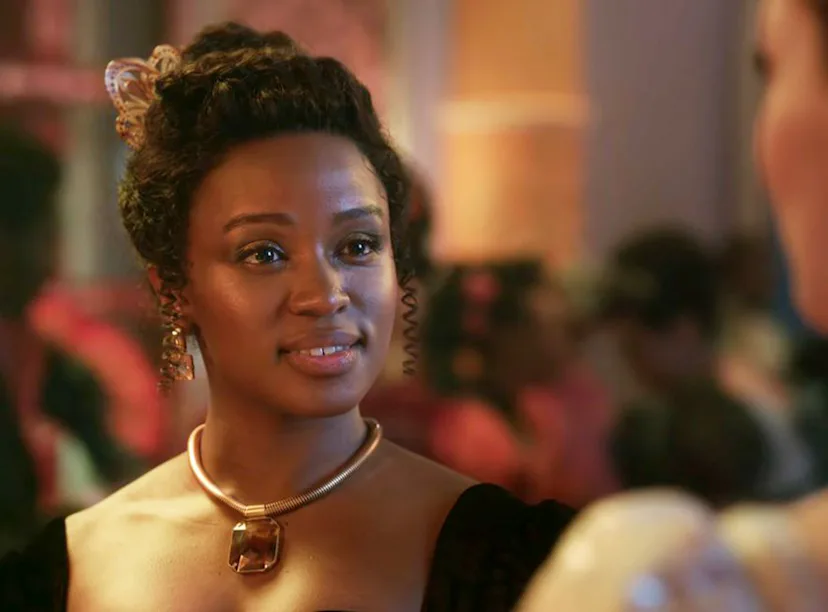 A woman with an elegant updo and a golden necklace gazes intently at someone in a warmly lit, bustli...