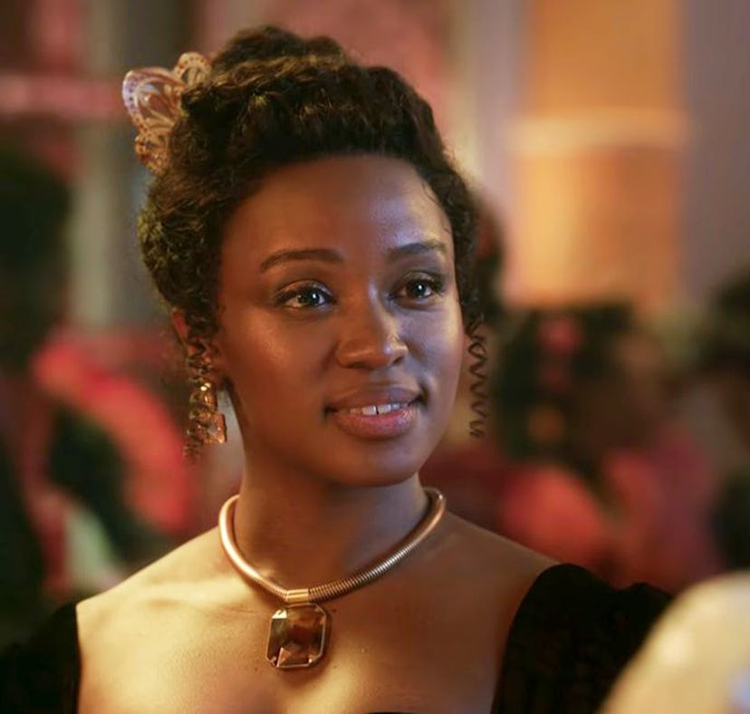 A woman with an elegant updo and a golden necklace gazes intently at someone in a warmly lit, bustli...