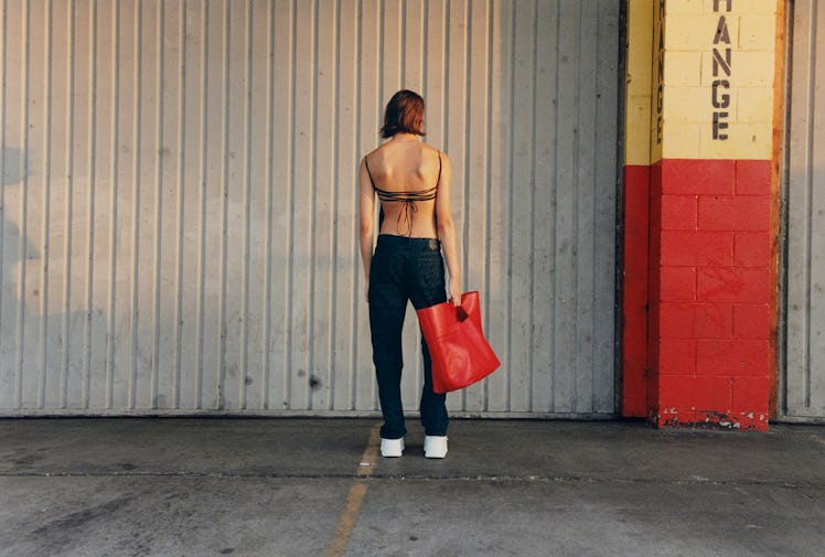 A person stands with their back to the camera, holding a red tote bag, in front of a grey wall with ...