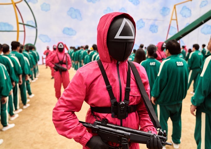 A person in a pink hooded suit with a geometric mask carries a rifle, standing among a crowd in green tracksuits.