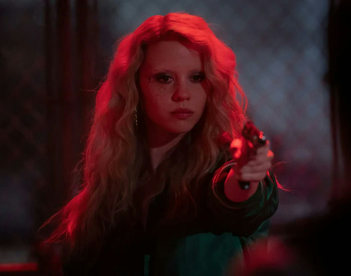 A woman with long blonde hair aiming a gun, intense expression, in a dimly lit, red-tinted room.