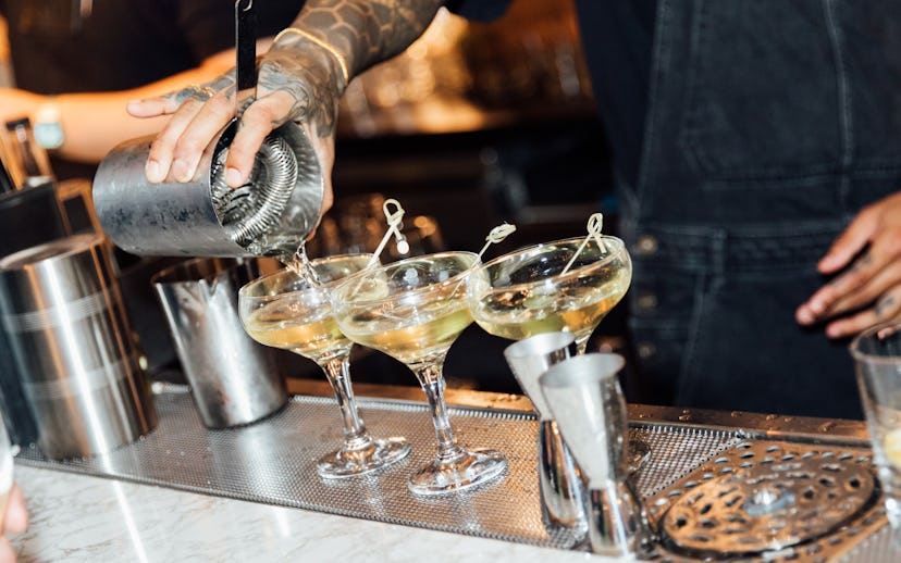 Bartender pouring drinks into three cocktail glasses on a bar counter, with cocktail-making tools ne...