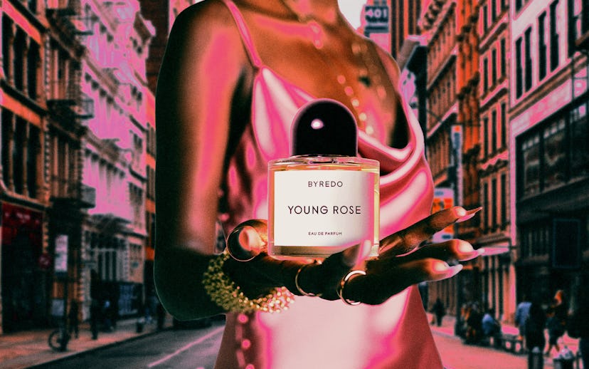 Woman in a pink dress holding a Byredo Young Rose perfume bottle with a blurry street background.