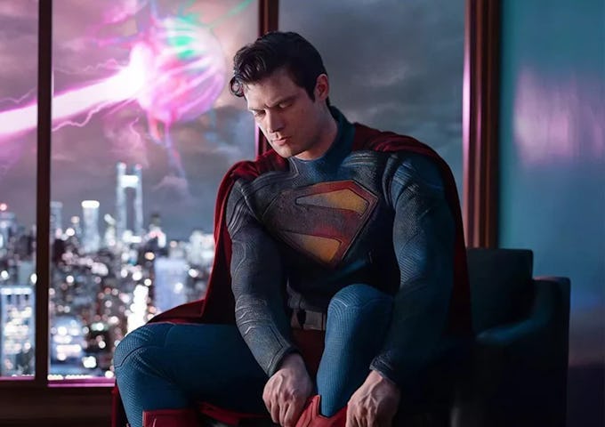 Superman in costume sits contemplatively beside a window overlooking a city skyline with a vibrant aurora in the sky.