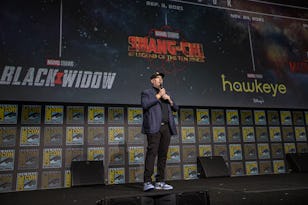 Man speaking at a podium at a Comic-Con event, with Marvel movie banners including "Black Widow" and "Shang-Chi" in the background.