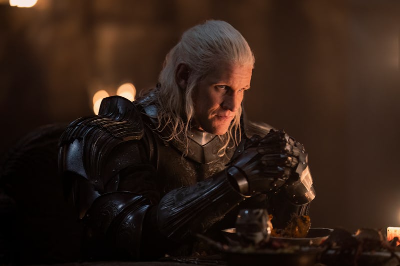 A knight with long white hair, wearing dark armor, sits at a dimly lit table intently gazing while h...