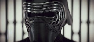 Close-up of a person wearing a dark, futuristic helmet with vertical, ribbed details, behind blurred metal bars.