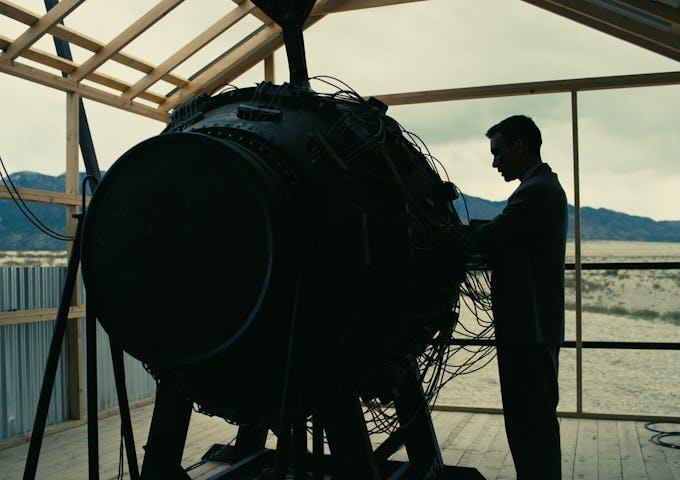 A man in silhouette works on a large, intricate machine inside a wooden structure, with a barren landscape visible in the background.
