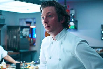 A chef with curly hair in a white chef's coat stands in a busy commercial kitchen, looking surprised...