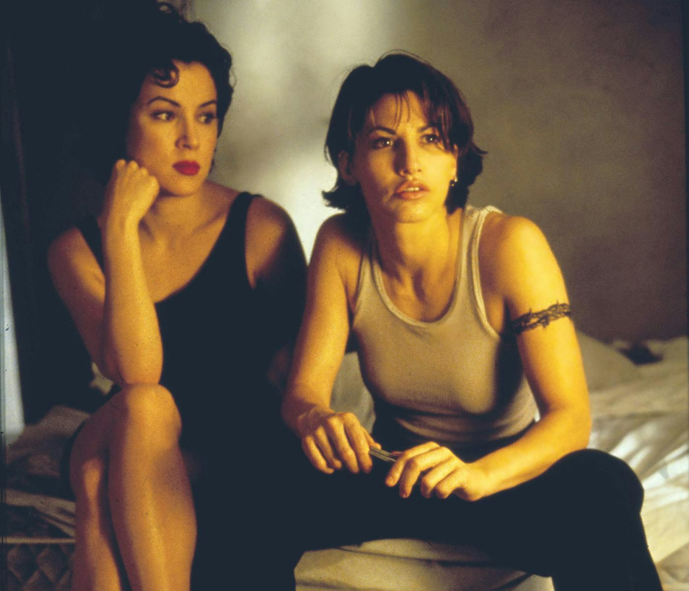Two women sitting on a bed, one in a black dress and the other in a tank top, looking thoughtful.