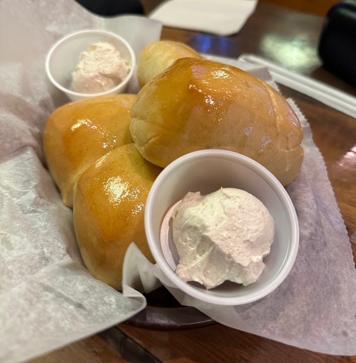 Three buttery Texas Roadhouse Rolls served on a paper liner with two small cups of whipped cinnamon ...
