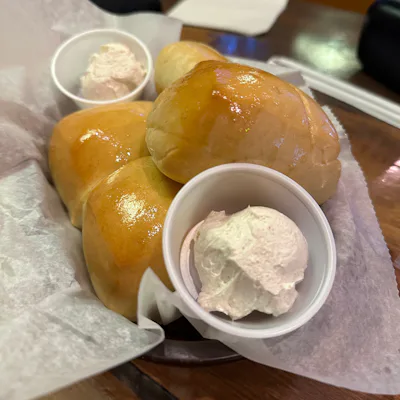 Three buttery Texas Roadhouse Rolls served on a paper liner with two small cups of whipped cinnamon butter sit on a wooden table.