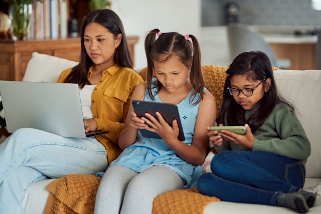 A mother and two young girls sit on a couch, each focused on their own device, in a cozy living room...
