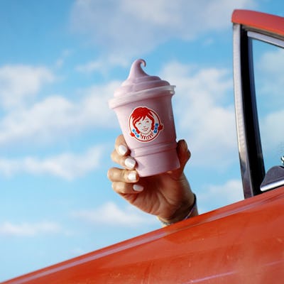 Hand holding a pink Wendy's frosty cup outside a car window against a blue sky.
