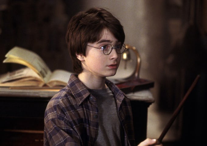 Young boy with glasses holding a wand, wearing a plaid shirt, surrounded by books in a dimly lit room.