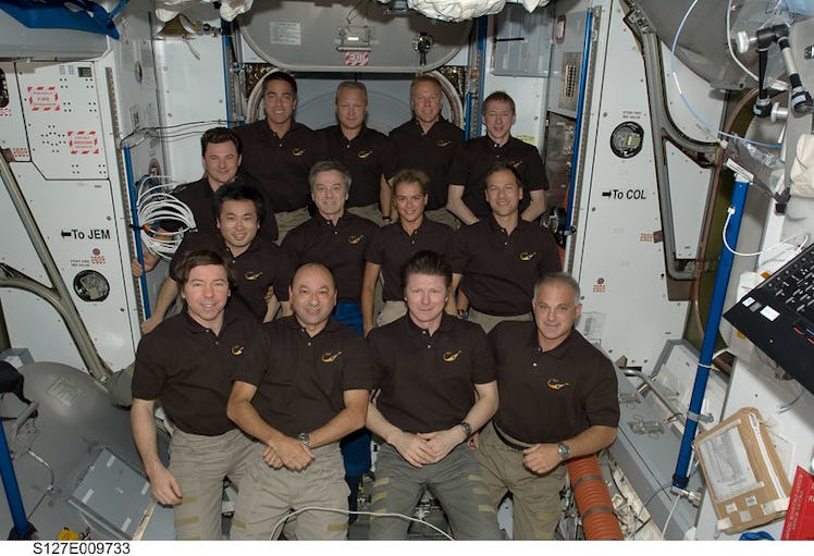 Thirteen astronauts, dressed alike in polos, pose together in three rows inside the space station. 