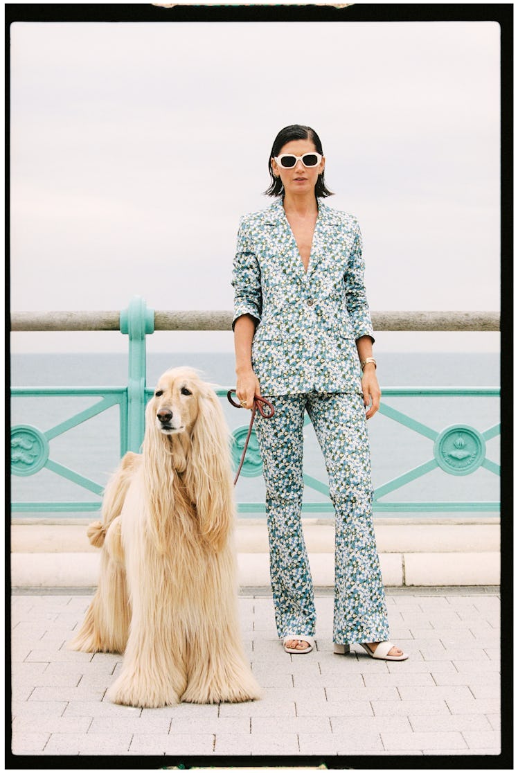 Woman in floral suit stands with a golden retriever by the seaside railing, both facing the camera.