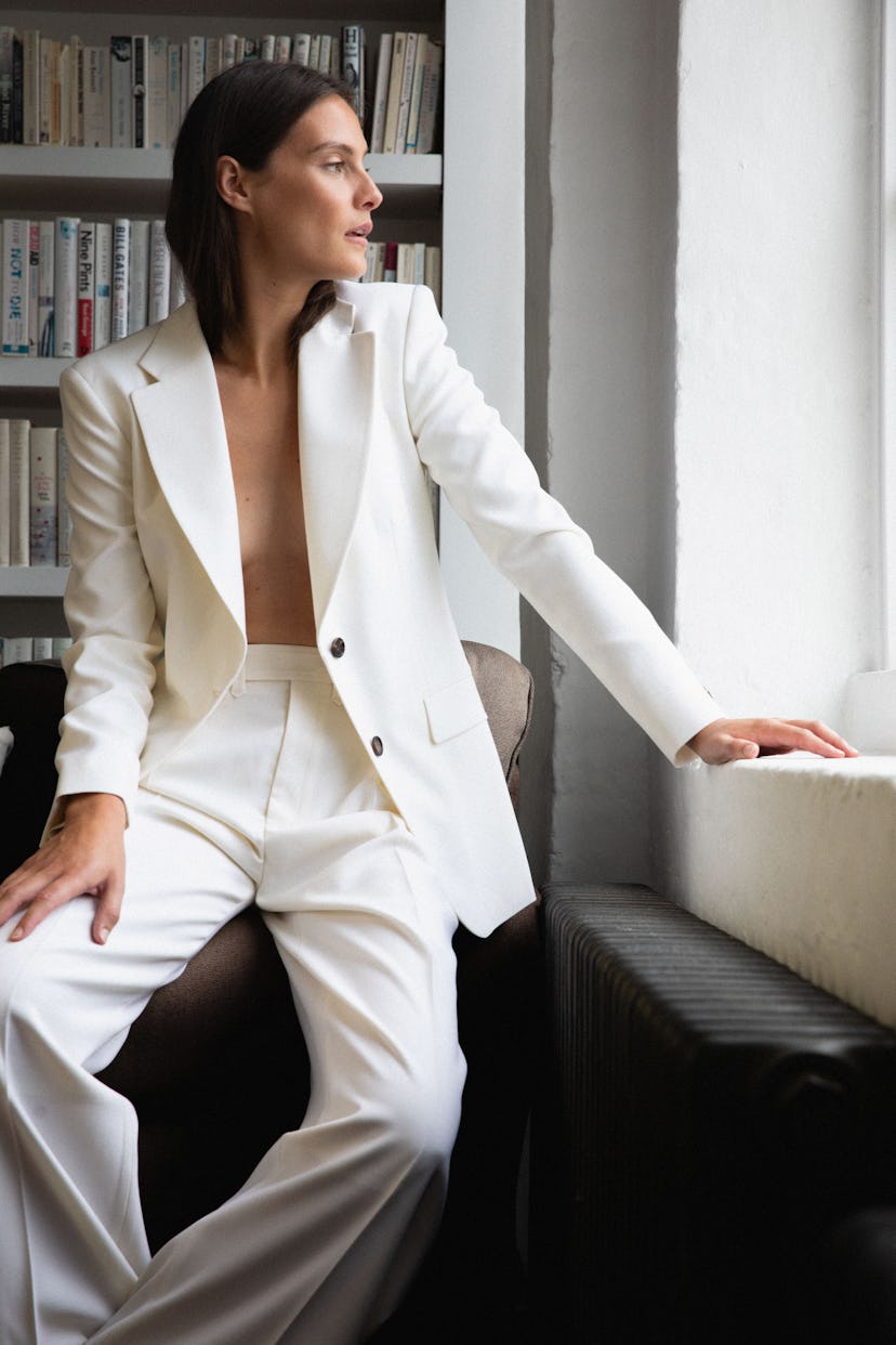 A woman in a stylish white suit sitting by a window, looking contemplatively to the side, with books...