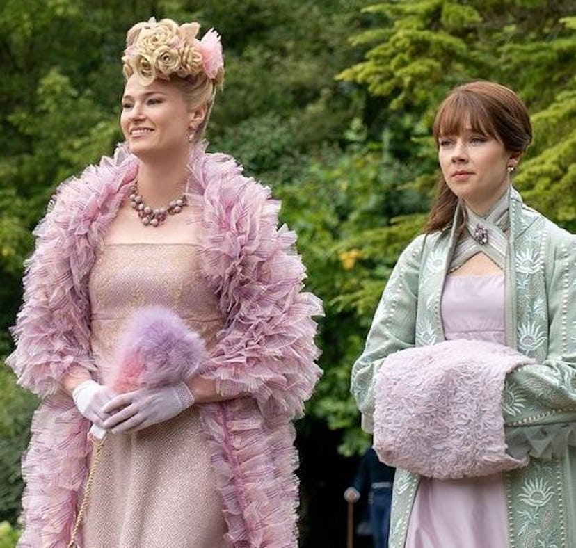 Two women in elaborate historical dresses and hats stand outdoors; one in pink with feathers, the ot...