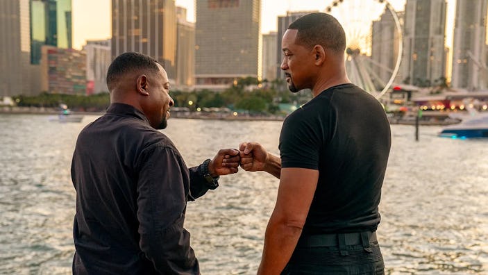 Two men fist-bumping on a waterfront with a cityscape behind them during sunset. They are engaged in...